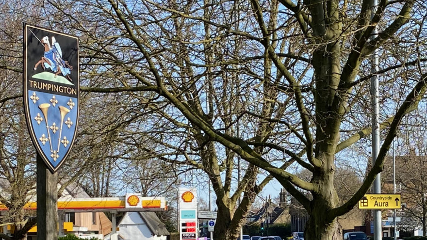 Trumpington shield shaped sign on the roadside displaying a knight riding a horse and two golden trumpets on a blue background